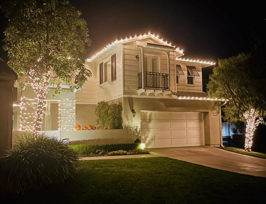 Two-story home outlined in warm-white Christmas lights, with illuminated trees beside its driveway