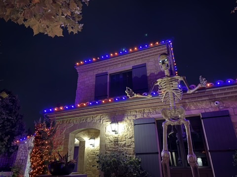 Large decorative skeleton in front of a stone home with orange and violet Halloween roofline lights