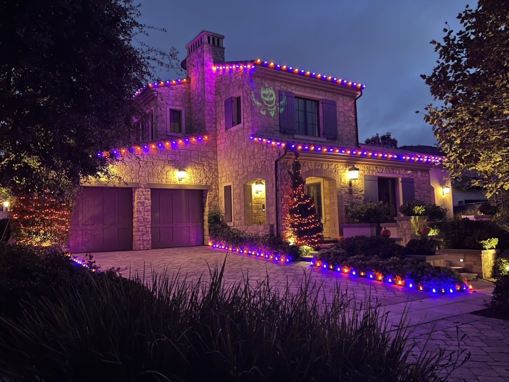 Stone home at dusk with orange and violet roofline lights, illuminated landscaping and a Halloween projection