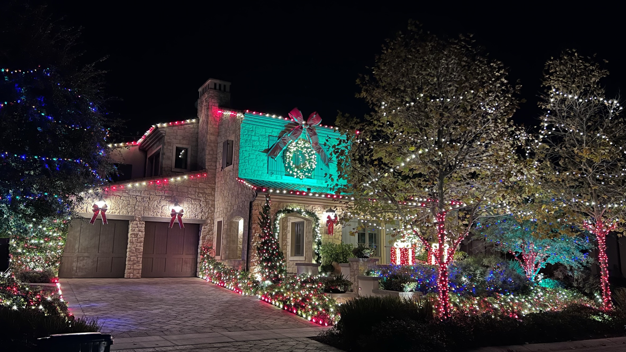 Stone home with red and white roofline lights, a wreath, and illuminated landscaping